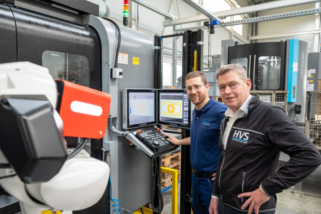 Thomas Schunn und Thomas Bär vor einem HURCO-Bearbeitungszentrum mit RoboJob-Roboterautomation